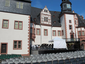 Eine Bühne vor der Mauer eines Renaissanceschlosses, auf der ein weißes Zelt steht. Davor Stuhlreihen.