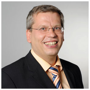 Roland Rüdinger lächelt herzlich zum Betrachter. Er hat ein Jackett und eine blauorangene Krawatte an.