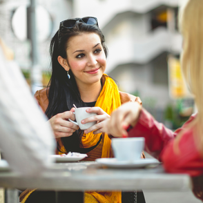 Frau sitzt entspannt in einem Café und genießt eine Tasse Kaffee, während sie sich mit einer Freundin unterhält.