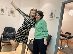 Im Bild - Margit Königsecker, Coach & Trainerin - sieht in die Kamera, bräunliches schulterlanges Haar, braunes Oberteil, im Hintergrund ein Bild