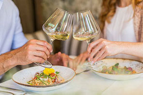 Ein Paar sitzt in einem Restaurant und stößt mit Weissweingläsern an. Auf dem Tisch stehen zwei Teller mit Essen. 
