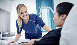Lächelnde Krankenschwester in blauer Uniform, die sich im Krankenzimmer zu einer Patientin hinunterbeugt, die in einem Bett liegt.