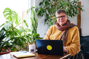 auf dem Foto zu sehen, ich sitze im Rollstuhl an einem kleinen Tisch, vor mir ein Laptop, ein Kalender und ein Kaffee, hinter mir jede Menge Zimmerpflanzen