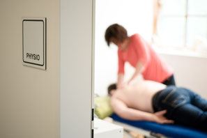 Blick durch eine Türe in einen Behandlungsraum, verschwommen sieht man eine Physiotherapeutin bei der Behandlung