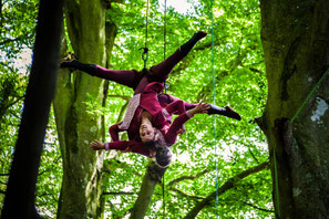 Artiste en pleine voltige aérienne, suspendue entre les arbres lors d'une performance de danse acrobatique en forêt, exprimant grâce et équilibre dans un cadre naturel.