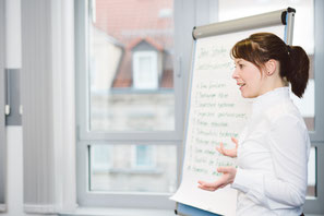 Ein Bild mit einer Frau, die in einem Konferenzraum erklärend auf ein Whiteboard zeigt