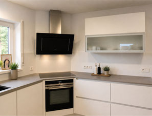 Grifflose Küche in Weiß matt mit grauer Arbeitsplatte, schwarzer Glas-Dunstabzugshaube und Glas-Hängeschrank von Holzdesign Rapp.