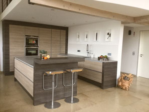 Grifflose Inselküche mit stirnseitigem Sitzplatz, raumhoher Hochschrankzeile in Eichedekor und grauer Dekton Arbeitsplatte