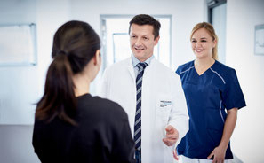 Ein Arzt und eine Krankenschwester begrüßen lächelnd eine Patientin in einem Krankenhausflur.