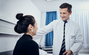 Ein Arzt in einem weißen Kittel mit Namensschild und Krawatte lächelt und spricht mit einer auf einem Bett sitzenden Patientin in einer Klinik.