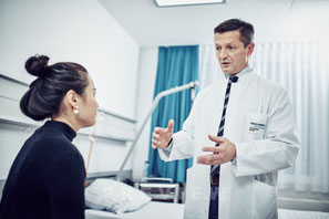 Ein Arzt in einem weißen Kittel mit Namensschild und Krawatte spricht erklärend mit einer Patientin in einem modernen Behandlungszimmer.