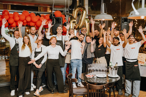 Mitarbeitende feiern gemeinsam das 10-jährige Jubiläum der L’Osteria Wien Hauptbahnhof. Rote Ballons, eine goldene „10“ und die fröhliche Pose der Gruppe unterstreichen die ausgelassene Stimmung.