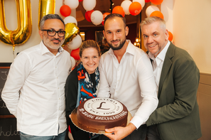 Vier Personen feiern das 10-jährige Jubiläum der L’Osteria in der Wiener Bräunerstraße und präsentieren gemeinsam eine festlich gestaltete Torte. Rot-weiße Ballons im Hintergrund unterstreichen die feierliche Stimmung.