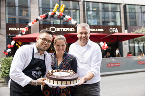 Drei Personen präsentieren vor der L’Osteria Innsbruck eine festlich gestaltete Torte anlässlich des 10-jährigen Jubiläums. Ballons und die Restaurantfassade im Hintergrund unterstreichen die feierliche Stimmung des Jubiläumstags.