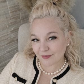 Portrait of a woman with blonde hair and black glasses in an elegant black-and-gold interior. She looks warmly into the camera, creating a natural and inviting atmosphere.
