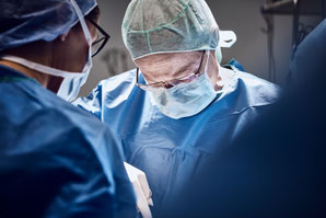 Chirurgisches Team, das in OP-Kleidung bei einer Operation konzentriert arbeitet, mit Fokus auf eine Person, die sich über den Patienten beugt.