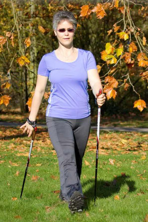 Trainerin läuft im Freien mit Nordic Walking-Stöcken auf einer grünen Wiese vor herbstlichen Bäumen. Man sieht eine akurate Ausführung eines typischen Schrittes mit Stock- und Armposition im Nordic Walking. Das Bild steht für Einzeltraining.