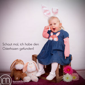 Ein 2-jähriges Mädchen mit einem Jeanskleid, auf dem ein Hase abgebildet ist, sitzt auf einer Holzkiste und hat einen Haarreif mit Hasenohren auf dem Kopf.