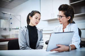 Ärztin in weißem Kittel zeigt einer Patientin Informationen auf einem Tablet.