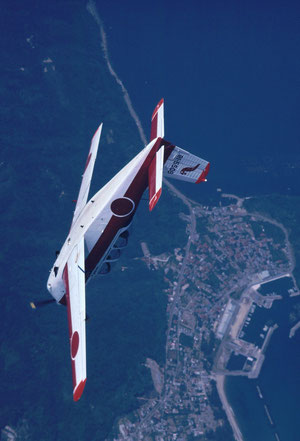 A JASDF cadet in a T-3 primary trainer does a loop over a coastal fishing town.