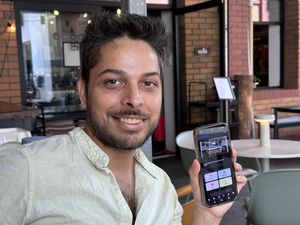 Junger Mann mit schwarzen Haaren und Bart, der ein aufgeknöpftes Hemd trägt, lächelt und zeigt sein Smartphone mit der DüsselQueers-App in die Kamera. Dahinter sind Café-Tische, eine Hauswand und Fenster zu sehen.