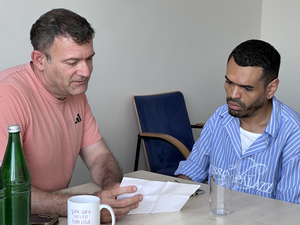 Zwei Männer sitzen an einem Tisch und unterhalten sich. Der Mann links trägt ein pinkes T-Shirt und hält ein Blatt Papier in der Hand. Der Mann rechts trägt ein blau-weiß gestreiftes Hemd.