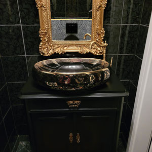 Antiker Waschtisch mit opulentem Becken in Gold und Schwarz mit barockem Spiegel mit Goldrahmen. Eine schwarze Toilette rundet das Bild ab.