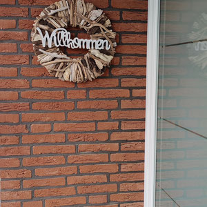Eingangsdeko am Hauseingang: Treibholzkranz mit Willkommen-Schild vor Klinkerwand, an einer weißen Haustür.