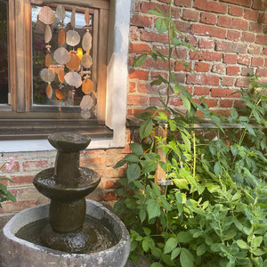 Capiz Windspiel in orange-apricot-natur am Wasserspiel in einem Naturgarten aufgehängt.