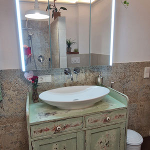 Nostalgischer Landhaus-Waschtisch in zarten Grün-/Brauntönen und einer aufgesetzten Waschschale. Darüber ein beleuchteter moderner Spiegel.