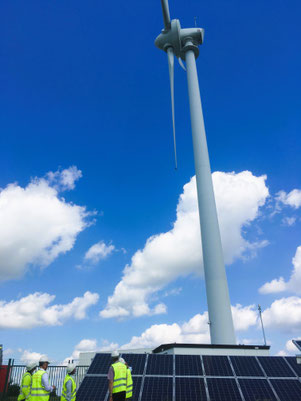 写真：太陽光パネルの前で作業する作業員と、背後にそびえる風力発電タービン。再生可能エネルギーの導入現場のイメージ