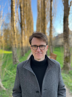 Das Bild zeigt Ulrich Rüdenauer, der mit grauem Mantel auf einer Allee steht. Er trägt kurze Haare und eine Brille.