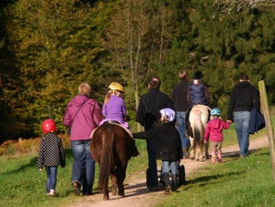 eine Gruppe mit Erwachsenen und kleinen Kindern läuft auf einem Weg in den Wald. Sie führen Ponys, auf denen Kinder sitzen