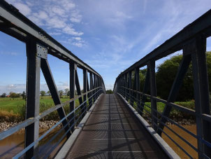 Das Foto ist auf einer Brücke aus einem Gitterrost stehend fotografiert. Man sieht, dass sie sehr schmal ist. Rechts und links ihre Stahlträger und Geländer, darunter der schlammbraune Fluss. Am anderen Ufer grüne Weiden und darüber der blaue Himmel. 