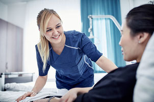 Lächelnde Krankenschwester in blauer Uniform, die sich im Krankenzimmer zu einer Patientin hinunterbeugt, die in einem Bett liegt.