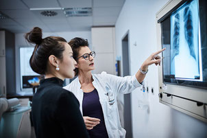 Eine Ärztin und ihre Patientin sind im Gespräch und blicken auf einen Leuchtkasten im Krankenhaus, in dem Röntgenaufnahmen gezeigt werden.