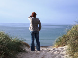 Ich blicke über die Nordsee. Man sieht mich von hinten zwischen 2 höheren, grasbewachsenen Dünen auf weißem Sand stehen. Meer und Himmel sind blau. Ich habe kurze, kastanienbraune Haare, trage Jeans und eine khakifarbene Bluse.