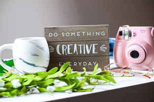 ピンク色のインスタントカメラとマグカップ。木製のボードにDO SOMETHING CREATIVE EVERYDAYのメッセージ。