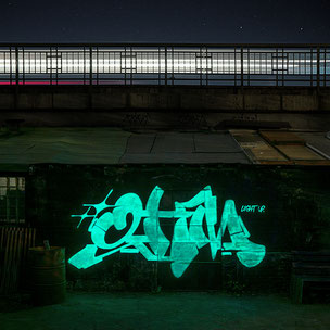 Eine Nachtaufnahme mit Sternen Himmel von der hamburger U-Bahn Trasse. Lichter eines vorbeifahrendes Zuges, unscharf durch die Bewegung und an der Wand der Trasse leuchtet ein Graffitischriftzug blau/grün auf.