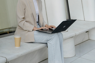 オフィスの休憩スペースのソファでノートパソコンを広げる女性。デニムにジャケット姿。傍らにテイクアウトコーヒーのプラカップ。