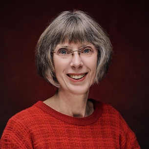 Een vrouw, Joke Zonneveld, staat voor een beukenhaag, grijzend ruimvallend haar, een roestkleurige sjaal en leren jas. warme haast groene ogen en een vriendelijke lach