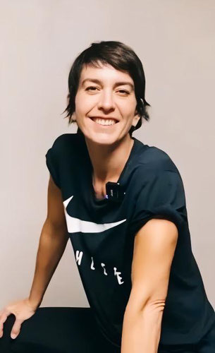Christiane Steiger ist in der Hocke, sie trägt dunkle, kurzarmige Sportbekleidung und lächelt vor einem rosafarbigem Hintergrund.