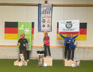 Drei Personen mit Hunden auf Podesten vor deutschen Flaggen, Banner mit Schrift 'dhv' und 'TEAM BLV' im Hintergrund.