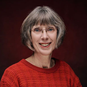 vriendlijk lachhende vrouw, Joke Zonneveld, met groen vest, lichtkrullend, en grijzend haar, met bril, groene ogen, voor beukenhaag