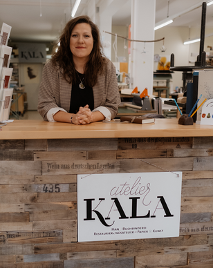 Profilbild von Katharina Lässig. Inhaberin vom atelier KALA. Die Frau hat gut schulterlanges braunes Haar und ist entspannt auf dem Empfangsthresen abgestützt. Sie lächelt freudig. Neben ihr liegt ein braunes Lederbuch.