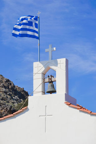 Weißer Kapellenturm mit Glocke vor einem Berg und wehender Griechenland Flagge
