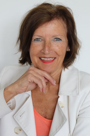 Margit Königsecker, Coach - Portrait, rechte Hand am Kinn, trägt weißen Blazer, orangenes T-Shirt