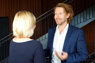 Coach Maximilian Daschner berät humorvoll eine Klientin während einer persönlichen Coaching-Sitzung in einem Gebäude