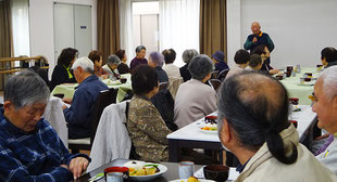 高齢者食事会で主催者のあいさつを聞く高齢者たち