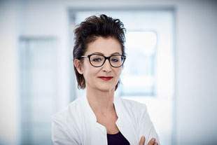 Frau Dr. med. Sabine Gailhofer, Oberärztin, im weißen Kittel, steht in einem modernen Klinikflur und schaut mit verschränkten Armen freundlich in die Kamera.